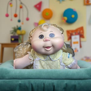 May include: A vintage doll with blonde curly hair, wearing a yellow dress with white lace trim. The doll has blue and brown eyes and is sitting in a teal-colored plush seat. The doll is a classic toy.
