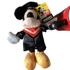 May include: A plush Mickey Mouse wearing a black graduation gown with a red and gold sash. He is holding a red megaphone with the word "Grad" on it. The tag on his arm says "Mickey The True Original 90 Years of Magic".