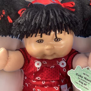 以下が含まれることがあります： 黒髪のツインテール、茶色の目、赤と白の模様のドレスを着たキャベツパッチキッズ人形。人形は「こんにちは！私はガビー・ナオンです。私の誕生日は8月30日です。私はクラフトをするのが好きです！」と書かれたタグを持っています。