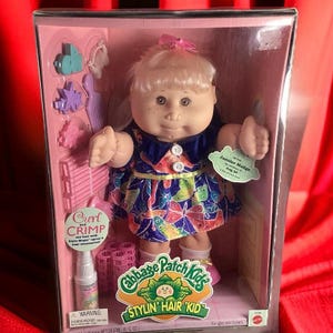 May include: Cabbage Patch Kids Stylin' Hair Kid doll with blonde hair, wearing a blue and purple butterfly print dress. The doll comes with hair styling accessories, including a bottle of hair spray, hair rollers, and a comb. The doll's name is Janalee Madge and she is age 50. The box is pink and white with the Cabbage Patch Kids logo.