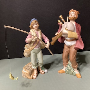 Könnte beinhalten: Zwei Porzellanfiguren, eine von einer Person, die eine Angelrute und einen Korb hält, die andere von einer Person, die Dudelsack spielt. Die Figuren sind in realistischen Farben bemalt und haben detaillierte Merkmale.