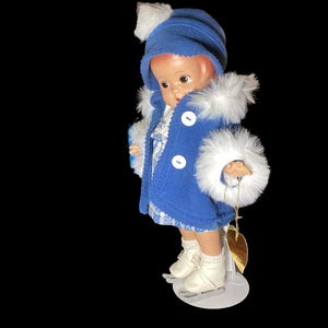 Op de afbeelding: Een porseleinen pop die een blauwe jas draagt met witte bontkraag en een blauwe hoed met een witte pompon. De pop draagt witte schaatsen en houdt een gouden hartbedel vast.