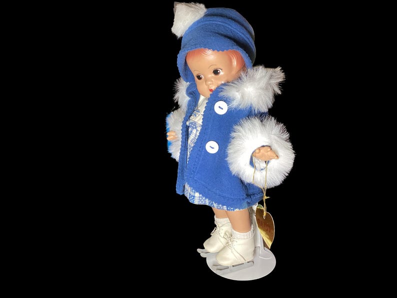 Op de afbeelding: Een porseleinen pop die een blauwe jas draagt met witte bontkraag en een blauwe hoed met een witte pompon. De pop draagt witte schaatsen en houdt een gouden hartbedel vast.