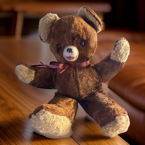 Può includere: Un orsacchiotto di peluche marrone con un nastro bordeaux intorno al collo. L'orso ha un viso bianco con occhi neri e un naso rosa. L'orso è seduto su una superficie di legno.