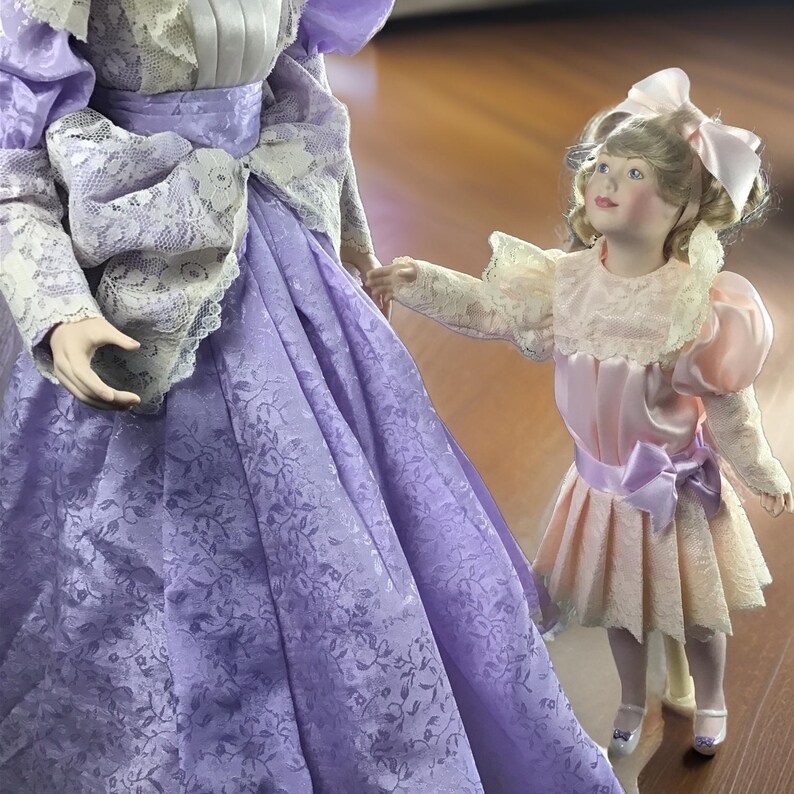 Puede incluir: Dos mu&ntilde;ecas de porcelana, una con un vestido lavanda con ribetes de encaje y la otra con un vestido rosa con una cinta lavanda. Las mu&ntilde;ecas est&aacute;n posadas como si estuvieran caminando juntas.