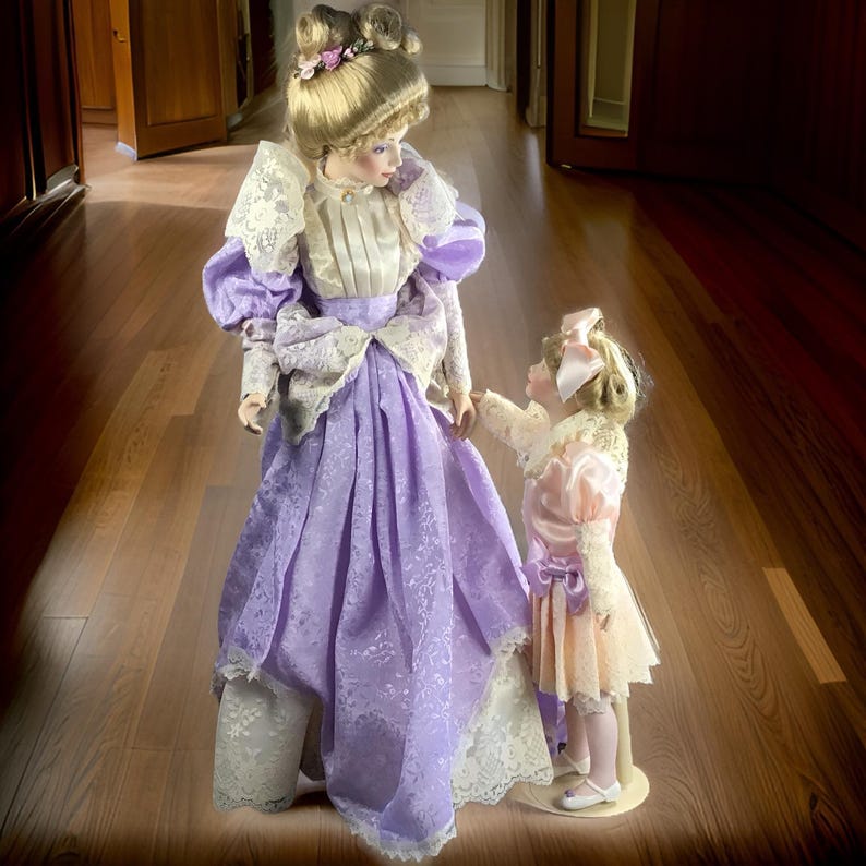 Puede incluir: Dos mu&ntilde;ecas de porcelana vestidas con ropa de inspiraci&oacute;n vintage. La mu&ntilde;eca m&aacute;s alta lleva un vestido lavanda con ribetes de encaje y una blusa blanca. La mu&ntilde;eca m&aacute;s peque&ntilde;a lleva un vestido rosa con ribetes de encaje y un lazo blanco. Ambas mu&ntilde;ecas tienen el pelo rubio y caras pintadas.