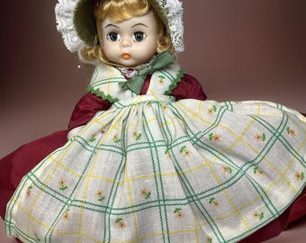May include: A vintage porcelain doll wearing a white dress with green and yellow plaid and floral accents. The doll has blonde hair, blue eyes, and a red bow. She is wearing a white lace bonnet.