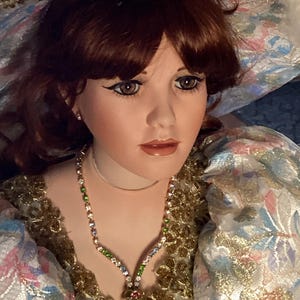 Könnte beinhalten: Porzellanpuppe mit rotbraunem Haar und braunen Augen. Die Puppe trägt ein farbenfrohes Kleid mit goldenen Verzierungen und eine Halskette mit Schmucksteinen. Der Hut der Puppe ist mit Gold und floralen Akzenten verziert. Die Puppe ist ein Sammlerstück.