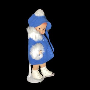Op de afbeelding: Een porseleinen pop die een blauwe jas draagt met witte bontkraag en een witte hoed. De pop draagt witte schaatsen en staat op een witte standaard.