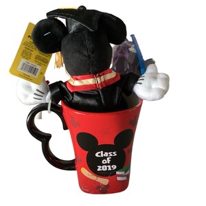 May include: A red ceramic mug featuring a black Mickey Mouse silhouette and the text "Class of 2019". A black plush Mickey Mouse wearing a graduation cap and gown is sitting inside the mug.