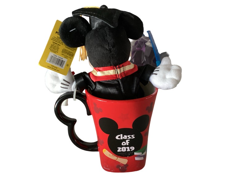 May include: A red ceramic mug featuring a black Mickey Mouse silhouette and the text "Class of 2019". A black plush Mickey Mouse wearing a graduation cap and gown is sitting inside the mug.