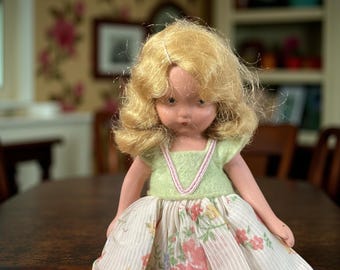 Puede incluir: Una muñeca vintage con cabello rubio y una parte superior verde con ribete rosa. La muñeca lleva una falda con estampado floral. La muñeca está sentada en una mesa marrón oscuro. La cara de la muñeca tiene una expresión seria.