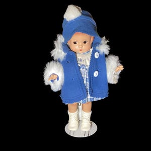 Op de afbeelding: Een blauwe en witte pop die een blauwe jas draagt met witte bontkraag en een witte hoed met een blauwe rand. De pop staat op een witte standaard.