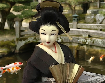 Puede incluir: Muñeca japonesa de geisha con kimono negro y abanico dorado. La muñeca tiene un peinado tradicional con horquillas decorativas. Peces koi nadan en el estanque de fondo, creando una escena serena y cultural.