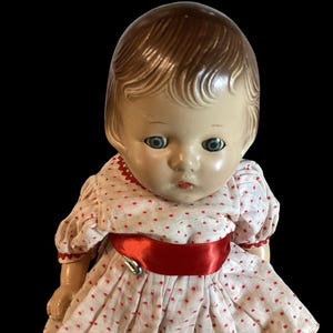 May include: A vintage porcelain doll with brown hair, blue eyes, and a white dress with red polka dots and a red satin sash.