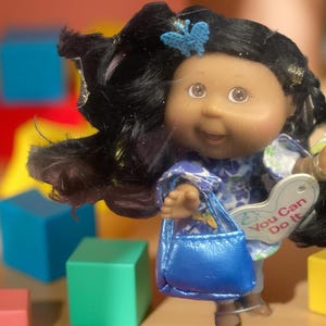Puede incluir: Una muñeca Cabbage Patch Kids con cabello negro y un bolso azul. La muñeca lleva un vestido floral y una pinza para el cabello con forma de mariposa azul. La muñeca sostiene un cartel que dice "You Can Do It".