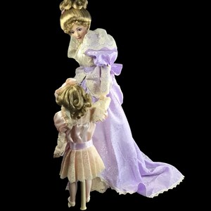 Puede incluir: Dos mu&ntilde;ecas de porcelana vestidas con vestidos de estilo vintage. La mu&ntilde;eca m&aacute;s alta lleva un vestido lavanda con ribetes de encaje y una superposici&oacute;n de encaje blanco. La mu&ntilde;eca m&aacute;s baja lleva un vestido rosa con ribetes de encaje y una superposici&oacute;n de encaje blanco. Ambas mu&ntilde;ecas tienen el pelo rubio y est&aacute;n posadas como si estuvieran caminando.