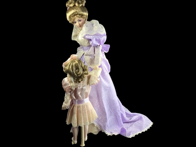 Puede incluir: Dos mu&ntilde;ecas de porcelana vestidas con vestidos de estilo vintage. La mu&ntilde;eca m&aacute;s alta lleva un vestido lavanda con ribetes de encaje y una superposici&oacute;n de encaje blanco. La mu&ntilde;eca m&aacute;s baja lleva un vestido rosa con ribetes de encaje y una superposici&oacute;n de encaje blanco. Ambas mu&ntilde;ecas tienen el pelo rubio y est&aacute;n posadas como si estuvieran caminando.