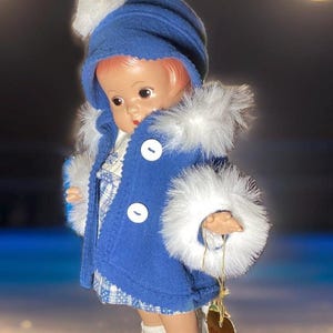 Op de afbeelding: Een porseleinen pop die een blauwe jas draagt met witte bontkraag en een witte hoed met een pompon. De pop draagt witte schaatsen en staat op een wit oppervlak. De pop heeft een label met de tekst "Daisy".