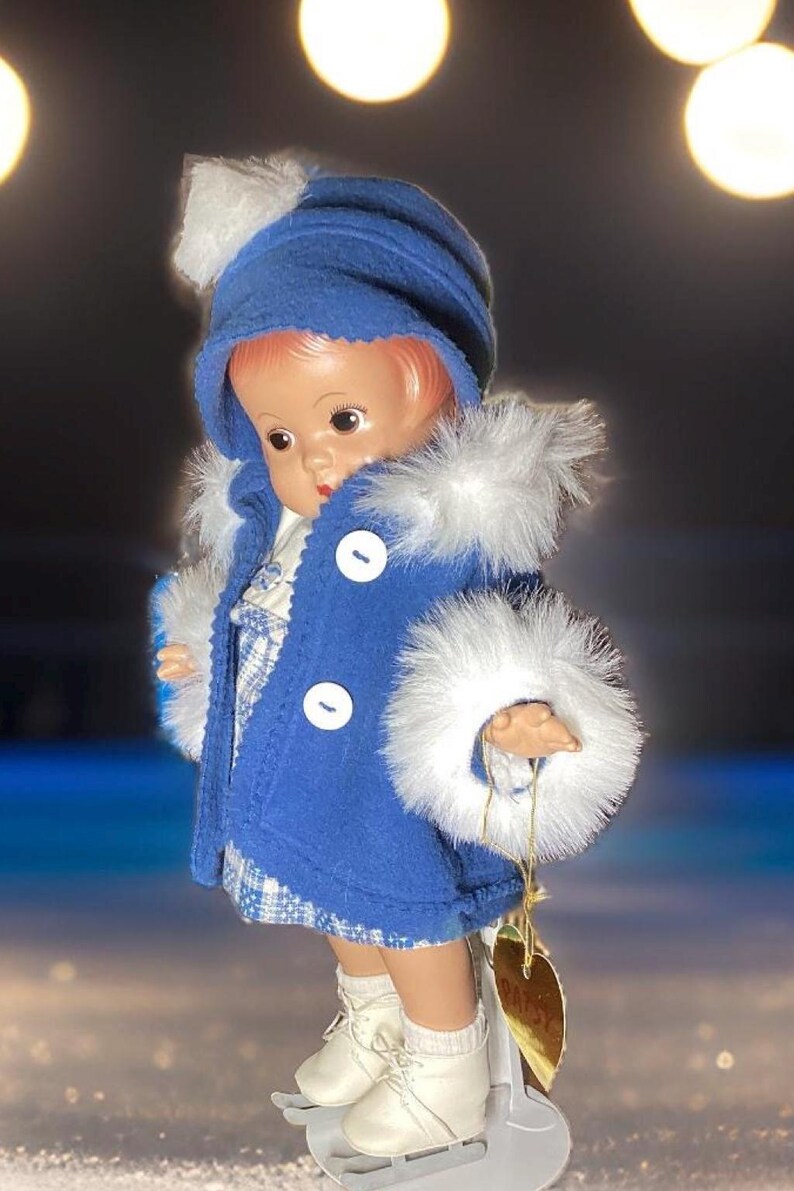 Op de afbeelding: Een porseleinen pop die een blauwe jas draagt met witte bontkraag en een witte hoed met een pompon. De pop draagt witte schaatsen en staat op een wit oppervlak. De pop heeft een label met de tekst "Daisy".
