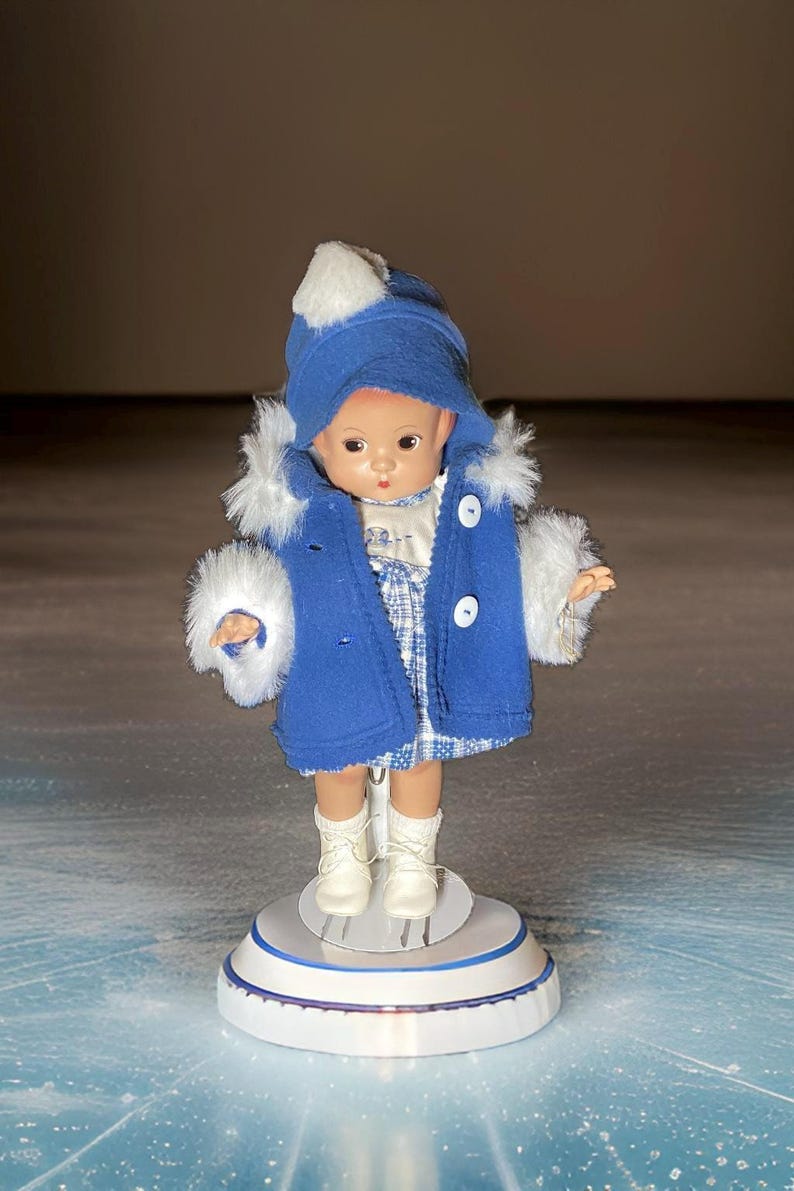 Op de afbeelding: Een porseleinen pop die een blauwe jas draagt met witte bontkraag en een blauwe hoed met een witte pompon. De pop staat op een wit platform met een blauwe rand.