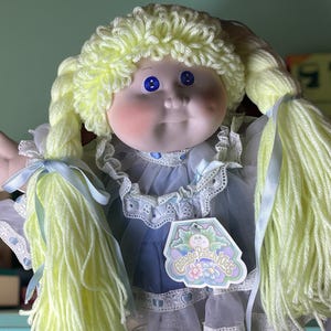 May include: A Cabbage Patch Kids doll with yellow yarn hair styled in pigtails. The doll has blue eyes, rosy cheeks, and is wearing a blue dress with white lace trim. A tag is attached to the dress.