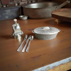May include: A miniature kitchen scene featuring a small metal pot with a lid, a fork, a knife, and a spoon. A metal clamp and a pan are also visible. The items are arranged on a brown wooden surface.