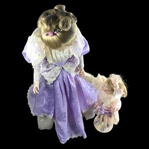 Puede incluir: Dos mu&ntilde;ecas de porcelana vestidas con vestidos morados y rosas con detalles de encaje. La mu&ntilde;eca m&aacute;s grande tiene el pelo rubio recogido en un mo&ntilde;o y sostiene la mano de la mu&ntilde;eca m&aacute;s peque&ntilde;a.