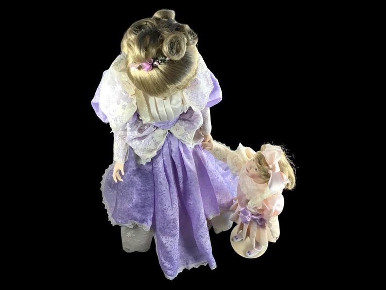 Puede incluir: Dos mu&ntilde;ecas de porcelana vestidas con vestidos morados y rosas con detalles de encaje. La mu&ntilde;eca m&aacute;s grande tiene el pelo rubio recogido en un mo&ntilde;o y sostiene la mano de la mu&ntilde;eca m&aacute;s peque&ntilde;a.