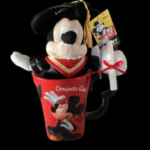 May include: A red ceramic mug with a black handle and a Mickey Mouse design. The mug says "Congrats Grad" and features a plush Mickey Mouse wearing a graduation cap and gown. The plush Mickey Mouse is sitting inside the mug.