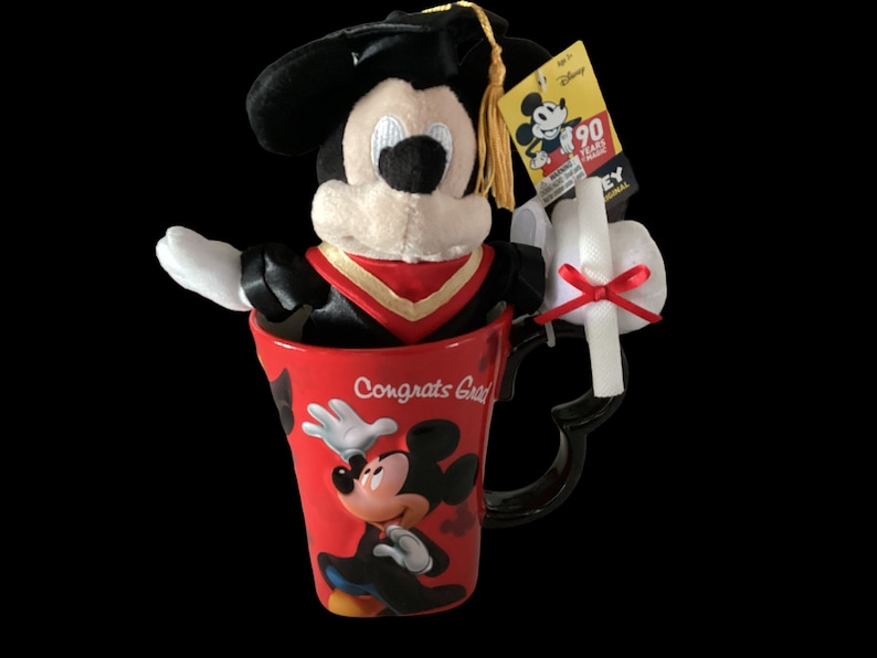 May include: A red ceramic mug with a black handle and a Mickey Mouse design. The mug says "Congrats Grad" and features a plush Mickey Mouse wearing a graduation cap and gown. The plush Mickey Mouse is sitting inside the mug.