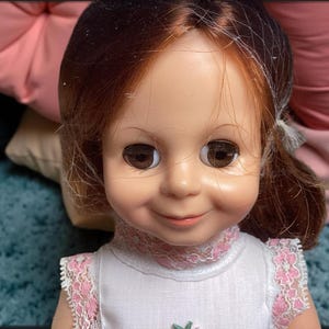 May include: A vintage doll with long brown hair, large blue eyes, and a sweet smile. She is wearing a white dress with pink lace trim.