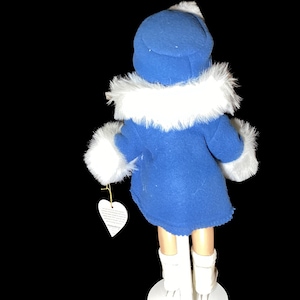 Op de afbeelding: Een porseleinen pop die een blauwe jas draagt met een witte bontkraag en witte laarzen. De pop houdt een wit hartvormig label vast.