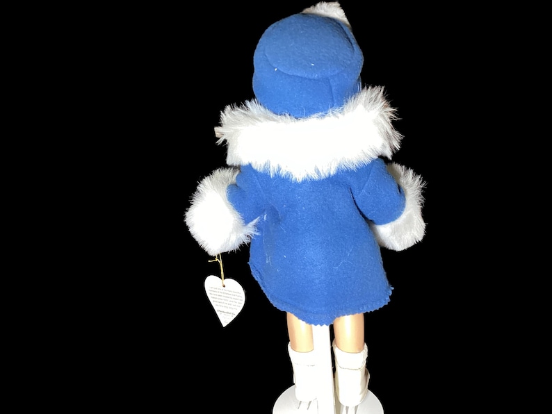 Op de afbeelding: Een porseleinen pop die een blauwe jas draagt met een witte bontkraag en witte laarzen. De pop houdt een wit hartvormig label vast.