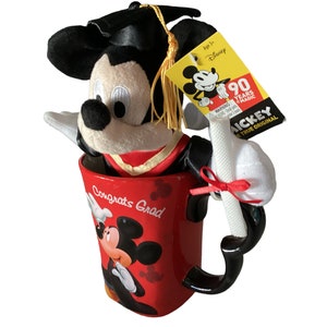 May include: Red ceramic mug with a graduation theme featuring Mickey Mouse. The mug has the words "Congrats Grad" on it. A plush Mickey Mouse wearing a graduation cap and gown is sitting inside the mug. The plush Mickey Mouse is holding a white scroll.
