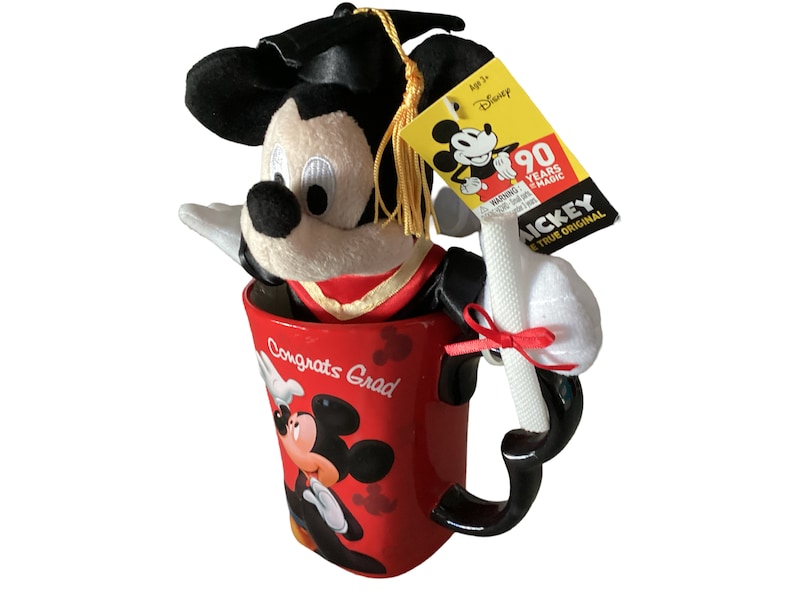 May include: Red ceramic mug with a graduation theme featuring Mickey Mouse. The mug has the words "Congrats Grad" on it. A plush Mickey Mouse wearing a graduation cap and gown is sitting inside the mug. The plush Mickey Mouse is holding a white scroll.