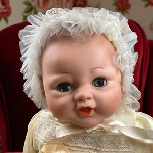 May include: A porcelain baby doll with a white lace bonnet and a white dress with lace trim. The doll has blue eyes and a painted smile.