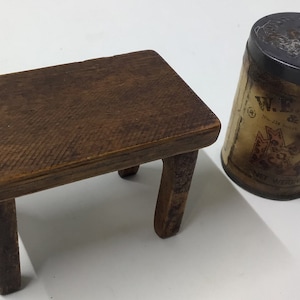 Puede incluir: Un pequeño taburete de madera antiguo con cuatro patas y una parte superior rectangular. El taburete está hecho de madera de color marrón oscuro. Junto a él hay una lata de metal vintage con una tapa oscura y una etiqueta descolorida.