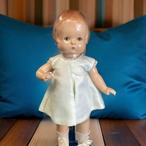 Op de afbeelding: Een vintage porseleinen pop met blond haar, die een lichtgroene jurk en witte sokken met strikjes draagt. De pop staat op een blauwe basis.