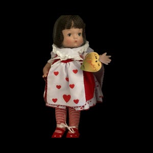 Puede incluir: Una muñeca de porcelana vestida como la Reina de Corazones de Alicia en el País de las Maravillas. Lleva un vestido blanco con corazones rojos, una falda a rayas rojas y blancas y zapatos rojos. La muñeca sostiene una etiqueta en forma de corazón que dice "Soy una  " con la palabra "Applause" escrita en ella.