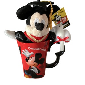 May include: A red ceramic mug with a black handle and a Mickey Mouse design. The mug says "Congrats Grad" and has a plush Mickey Mouse wearing a graduation cap and gown inside. The plush Mickey Mouse is holding a white diploma with a red ribbon.