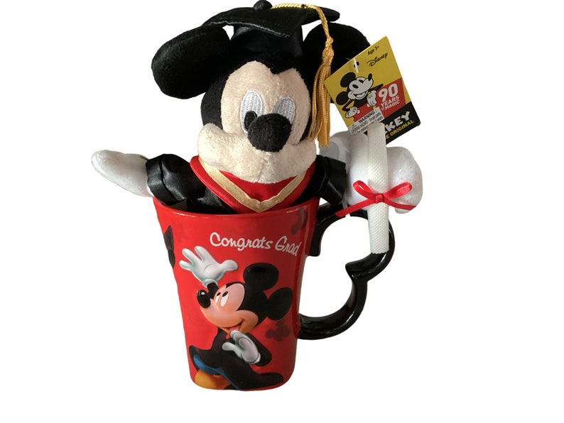 May include: A red ceramic mug with a black handle and a Mickey Mouse design. The mug says "Congrats Grad" and has a plush Mickey Mouse wearing a graduation cap and gown inside. The plush Mickey Mouse is holding a white diploma with a red ribbon.