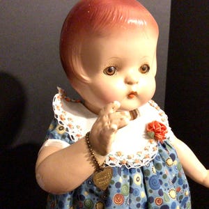 Puede incluir: Una muñeca vintage con rostro pintado, cabello rojo y ojos marrones. La muñeca viste un vestido azul con un babero blanco y una flor roja. Una pulsera de abalorios dorada está en el brazo de la muñeca.