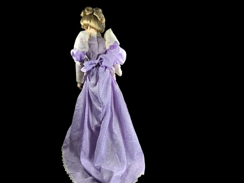 Puede incluir: Una mu&ntilde;eca de porcelana con un vestido lavanda con ribete de encaje blanco y un lazo en la cintura.