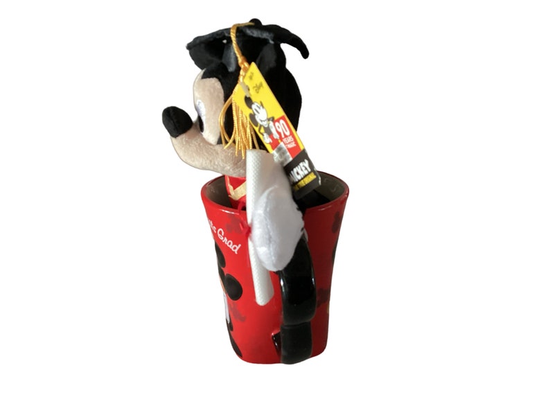 May include: A red ceramic mug with a black handle and a Mickey Mouse design. The mug says "It's Grad" and has black polka dots. A plush Mickey Mouse wearing a graduation cap and gown is sitting inside the mug. The plush Mickey Mouse has a yellow tag that says "Mickey 90 Years of Magic".
