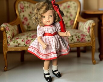 May include: A vintage doll with curly blonde hair, green eyes, and a red bow. The doll wears a white dress with red polka dots, a red belt, and black shoes with white socks. The doll is standing in front of a floral patterned chair.