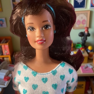 May include: A brunette doll with blue eyes and a white shirt with a turquoise heart pattern. The doll is wearing a blue headband and blue earrings.