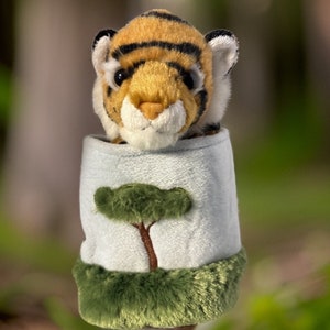 May include: A plush tiger toy peeking out of a blue and green fabric pouch with a green tree design. The pouch has a fuzzy green trim.
