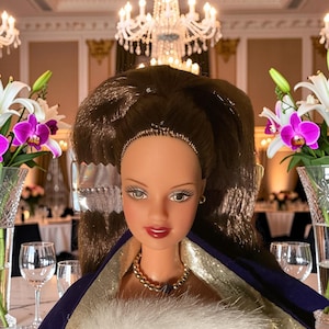 May include: A doll with brown hair and a gold necklace, wearing a purple and gold outfit with a white fur trim. The doll is in a formal setting with chandeliers, flowers, and wine glasses.