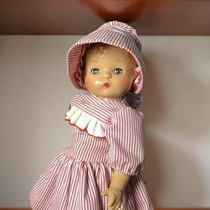 Puede incluir: Muñeca vintage con un vestido a rayas rojas y blancas y un gorro a juego. La muñeca tiene ojos verdes, mejillas rosadas y labios rojos. El vestido presenta un ribete de volantes blancos con bordes rojos.